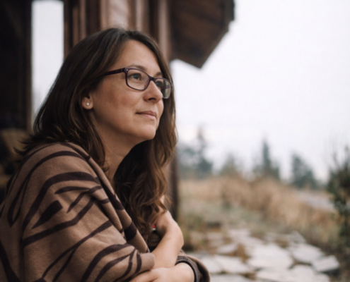 Nachdenkliche Frau sitzt vor einem rustikalen Holzhaus und blickt in die Ferne – symbolisch für innere Reflexion und das Lösen von Konflikten